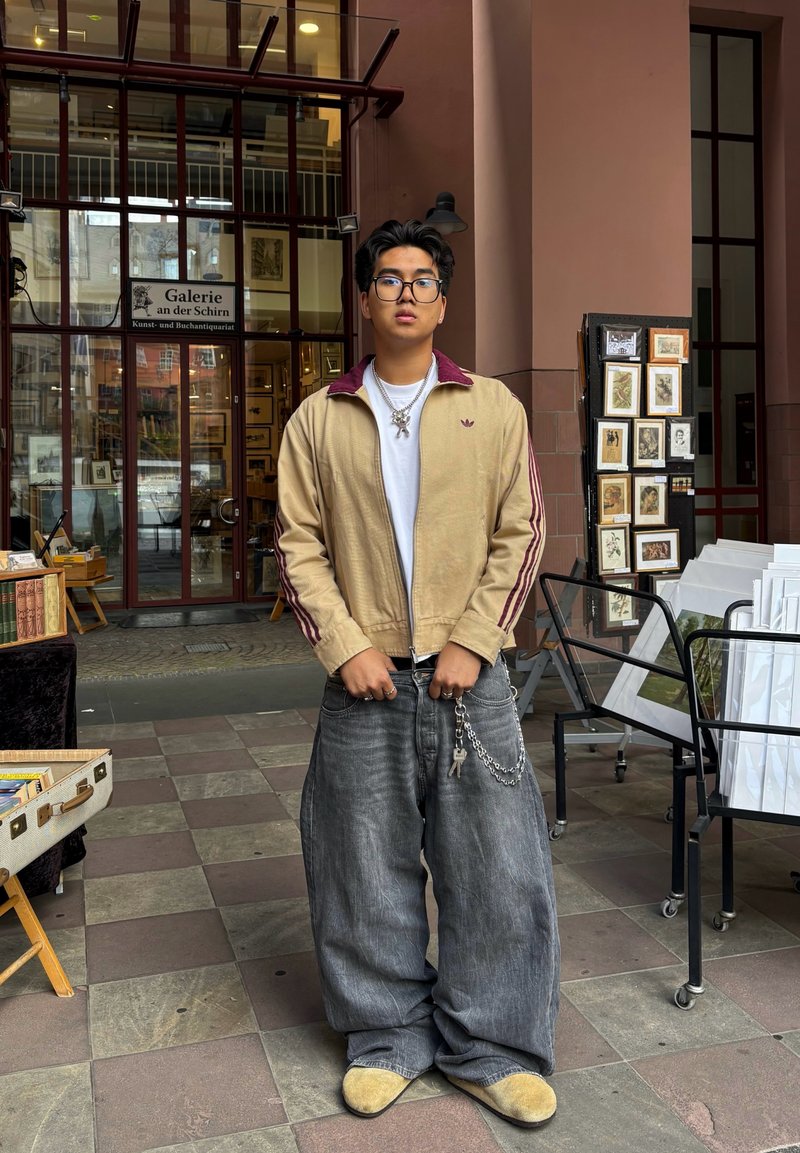 Junge Person mit Brille, die eine beigefarbene Jacke und weite Jeans trägt, steht vor dem Eingang der Galerie an der Schirn mit gerahmten Kunstwerken in der Nähe.