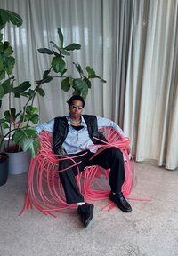 Sedia in plastica flessibile di colore rosa con un design a spirale irregolare. Un modello vestito con un gilet nero, una camicia blu e scarpe nere è seduto tra la vegetazione.