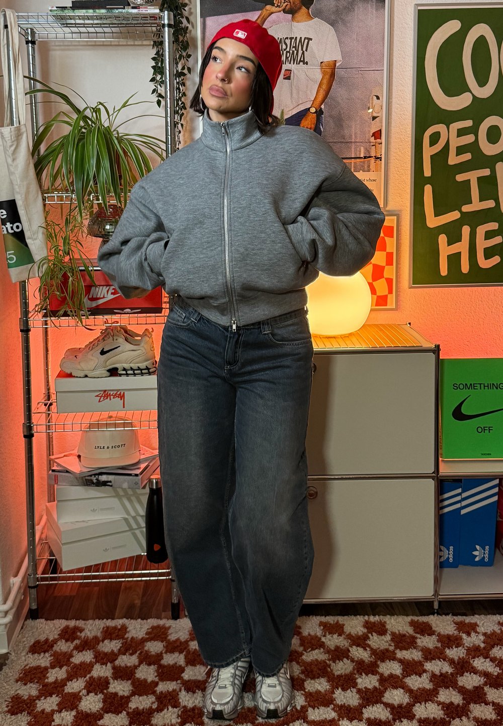 Siva jopa z zadrgo, modre široke kavbojke in srebrni supergi. Rdeča kapa doda barvni poudarek. V ozadju so police z barvitimi škatlami in rastlinami.
