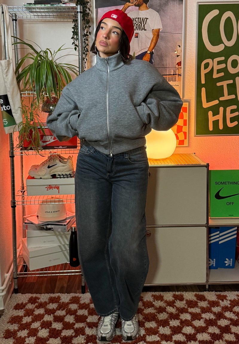 Sweat-shirt gris à fermeture éclair, jeans bleus amples et baskets argentées. Casquette rouge pour une touche de couleur. En arrière-plan, des étagères avec des boîtes colorées et des plantes.