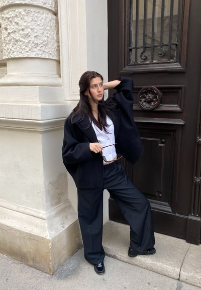 Chaqueta negra oversize, blusa blanca con botones, pantalones negros anchos, de pie contra una pared blanca texturizada y una puerta marrón, sosteniendo unas gafas.