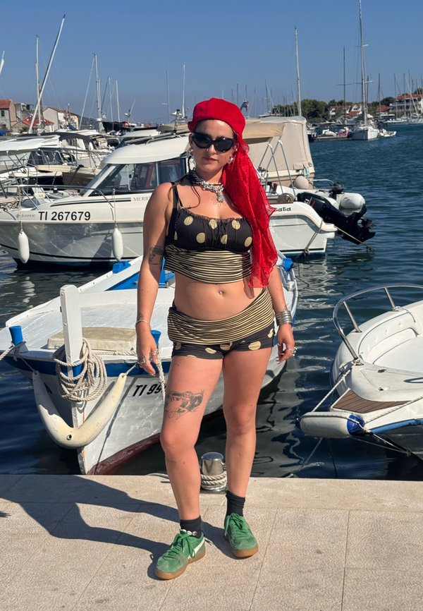 Persona con top nero a pois dorati e pantaloncini, fazzoletto rosso in testa, scarpe verdi, in piedi su un molo con barche e acqua sullo sfondo.