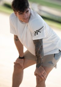 Witte T-shirt met korte mouwen, zwarte accenten en gestippelde patronen, gedragen met lichtbeige shorts. Tatoeages zichtbaar op de linkerarm; handen op de knieën.