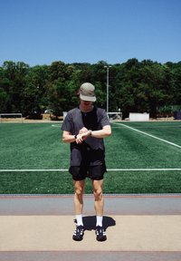 Sportóra, fekete rövidujjú póló, fekete sortok, fehér zoknik, fekete sportcipők fehér részletekkel; zöld műfű és kék ég háttér.