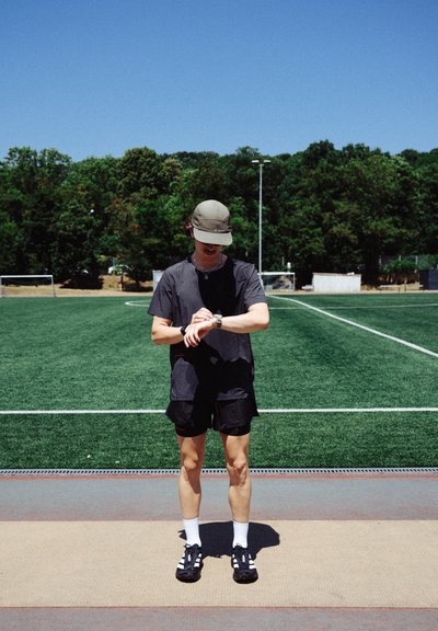 Reloj deportivo, camiseta negra de manga corta, pantalones cortos negros, calcetines blancos, zapatillas negras con detalles blancos; fondo de césped verde y cielo azul.
