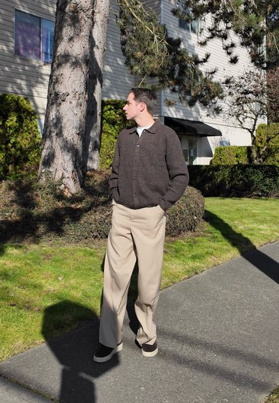 Hombre con chaqueta marrón y pantalones beige de pie en la acera, manos en los bolsillos, mirando hacia su izquierda cerca de árboles y arbustos junto a una casa.