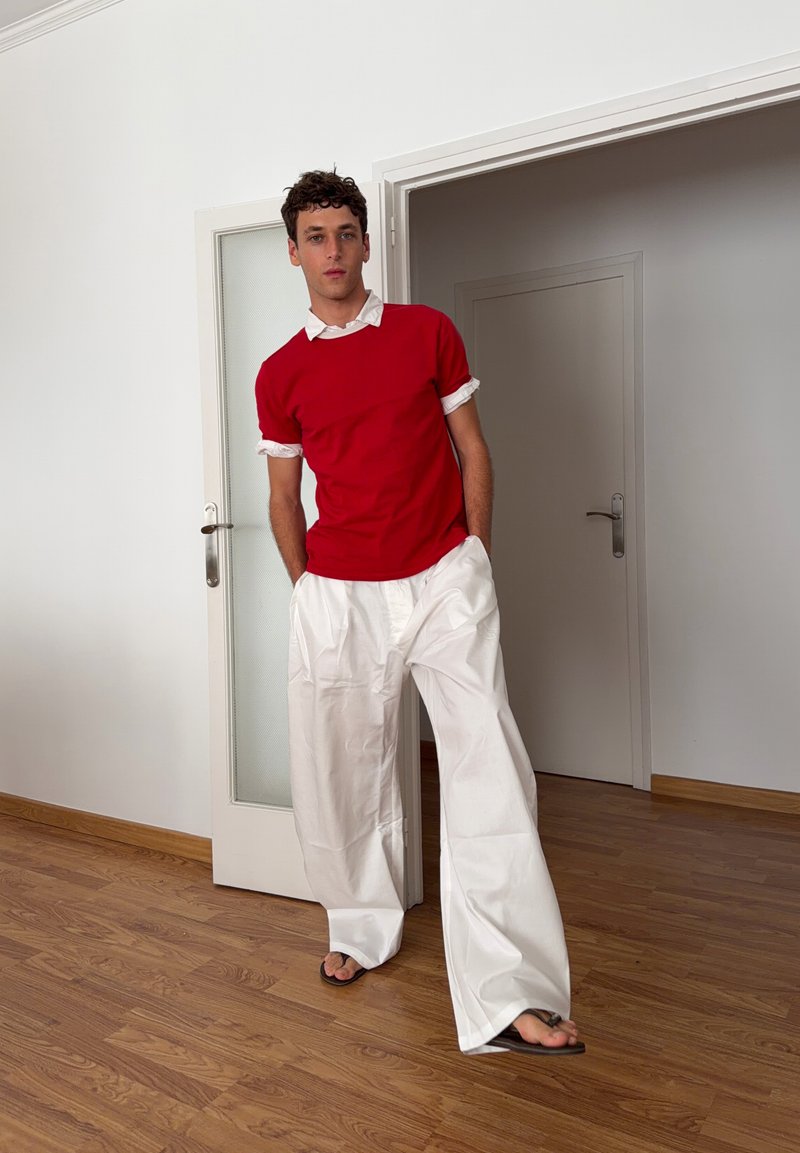 Homme debout à l'intérieur portant un pull rouge à manches courtes sur une chemise blanche, large pantalon blanc et sandales, sur un sol en bois près d'une porte.