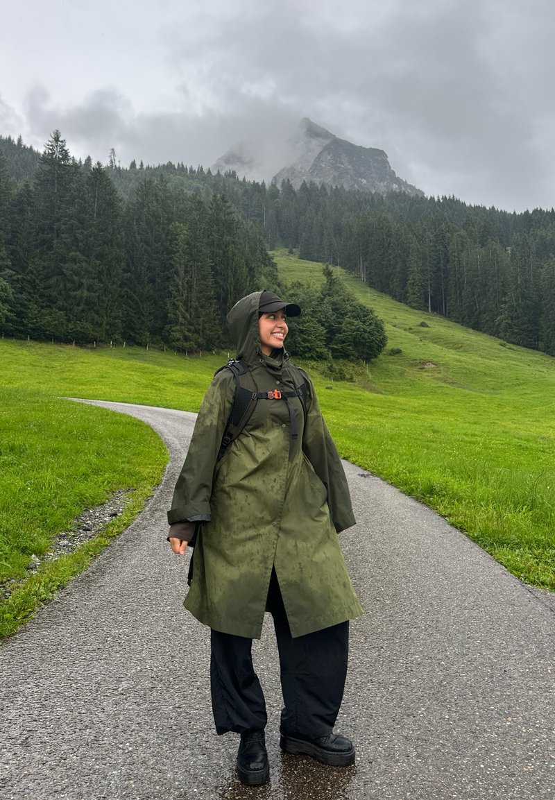 Giacca impermeabile verde, pantaloni neri e scarpe nere pesanti. Il paesaggio include alberi e montagne sotto un cielo nuvoloso. La strada bagnata curva.