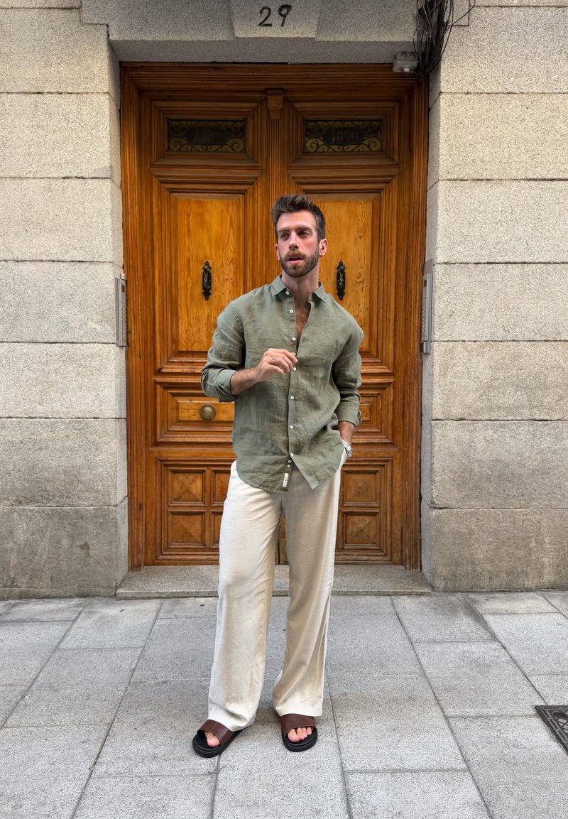 Homme en chemise verte et pantalon beige debout sur le trottoir devant une grande porte en bois numérotée 29, avec une main dans la poche et l'autre levée.