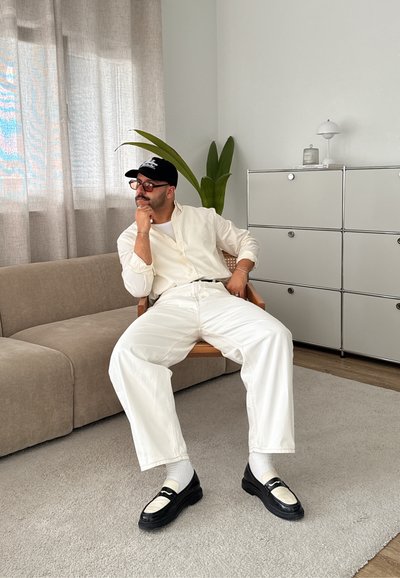 Camisa blanca de manga larga, jeans blancos, mocasines negros con acentos blancos, sentado en un sofá beige, alfombra gris claro, planta verde cercana.