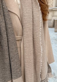 Κοντινό πλάνο ενός ατόμου που φορά στρωματώδη μπεζ και γκρι μάλλινα παλτά και κασκόλ, με σγουρά κόκκινα μαλλιά να είναι ορατά σε ένα χιονισμένο υπαίθριο τοπίο.