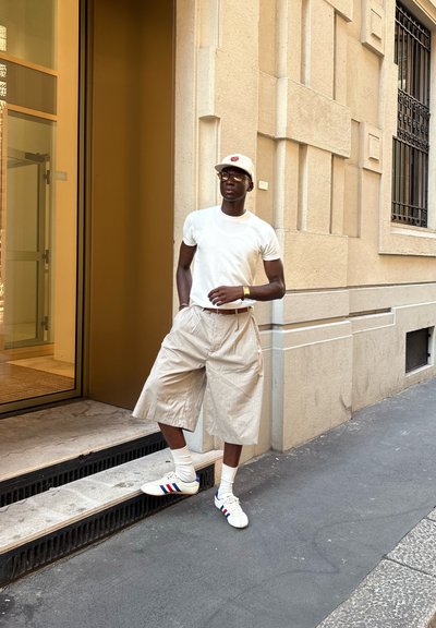 Pantalones cortos a rayas beige, camiseta blanca y zapatillas blancas con rayas azules y rojas. Los accesorios incluyen una gorra de color claro y un reloj.
