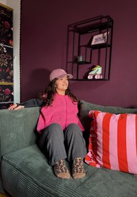Femme en gilet rose et casquette assise sur un canapé vert avec un coussin rayé rouge et rose, mur violet derrière avec une étagère géométrique noire et un patin à roulettes.