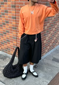 Een oranje lange mouwen shirt, zwarte losse shorts, witte sokken, zwart-wit schoenen en een zwarte gewatteerde tas met een gevarieerde textuur.