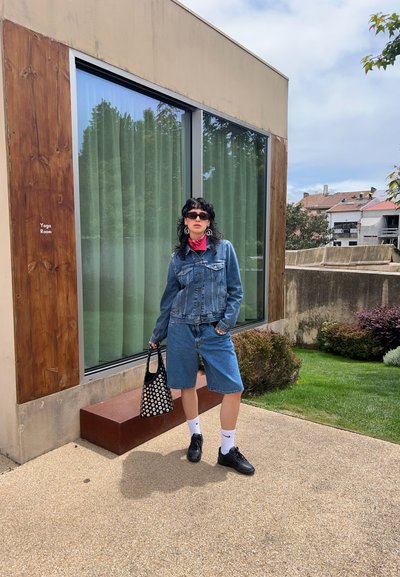 Combinación de chaqueta de denim y pantalones cortos; zapatillas negras; calcetines blancos; bandana roja; bolsa negra con patrones; cortinas de ventana verdes al fondo.