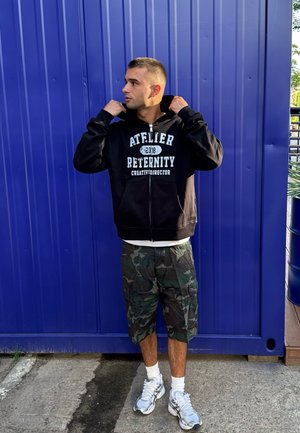 Sudadera negra con cremallera y texto blanco, pantalones cortos de camuflaje y zapatillas grises. De pie sobre el pavimento frente a un fondo azul. Atuendo casual.
