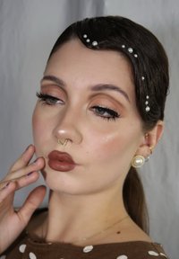 In primo piano, un volto con rossetto marrone chiaro, anello nasale dorato e accenti di perle nei capelli. La pelle ha una leggera lucentezza e gli occhi sono accentuati con eyeliner.