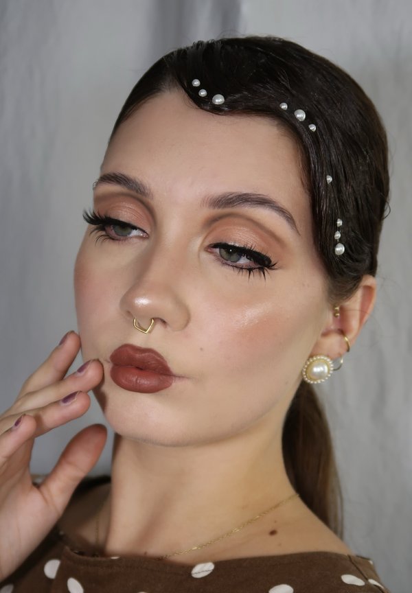 Close-up of a face with light brown lipstick, gold nose ring, and pearl hair accents. Skin has a soft sheen, and eyes are accentuated with eyeliner.