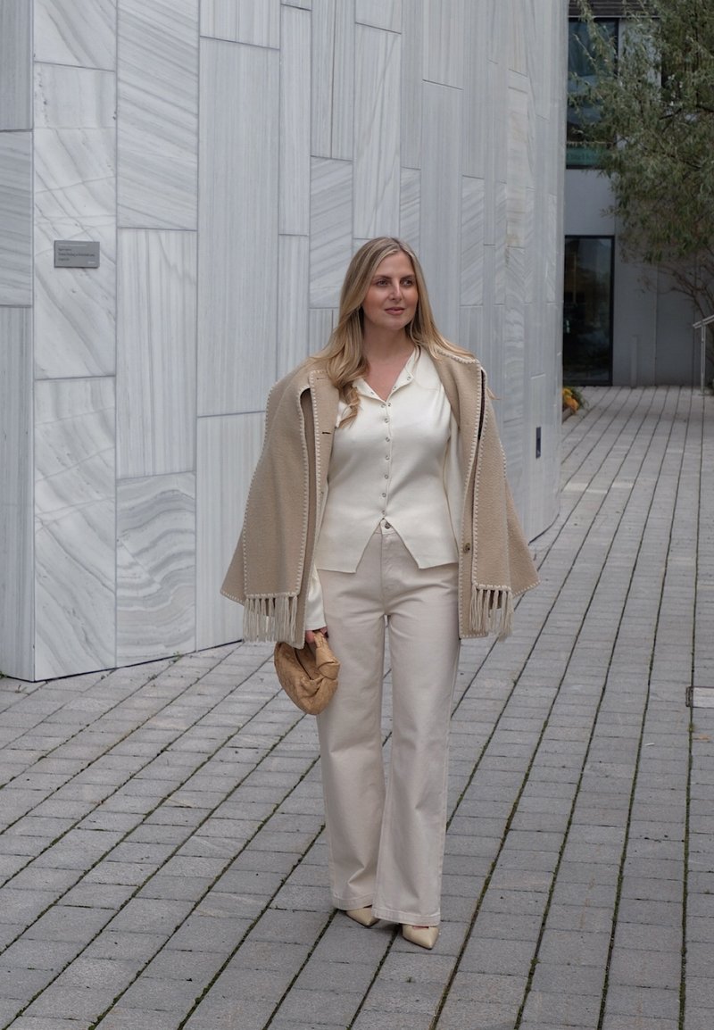 Beiger Poncho mit Fransen, cremefarbene Bluse und hellbeige ausgestellte Hose. Hält eine beigefarbene Clutch, läuft auf einem Steinpflaster vor einer Marmorwand.