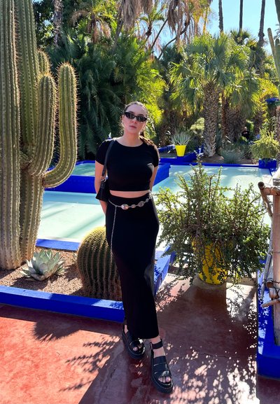 Top corto negro con mangas cortas, falda maxi negra, sandalias gruesas negras, cinturón de cadena plateado, rodeada de cactus y macetas coloridas.