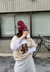 Personne portant un bonnet bordeaux et des lunettes, buvant dans une tasse, debout sur une rue pavée près d'un bâtiment avec un vélo garé à proximité.