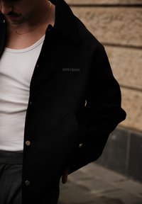 Svart jakke med trykknapper i teksturert stoff med brystlomme som har en diskré logo. Designet med krage og løs passform.