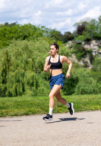 Sujetadora deportiva negra, pantalones cortos azul marino y zapatillas deportivas negras con suelas blancas. Corriendo por un sendero rodeado de vegetación y un cielo azul de fondo.