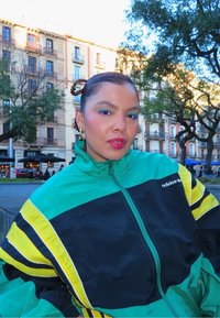 Femme aux cheveux tressés et boucles d'oreilles en anneau doré portant une veste Adidas verte, noire et jaune, posant en plein air avec des bâtiments et des arbres en arrière-plan.