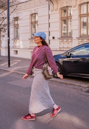 Mujer con camiseta de rayas, falda de mezclilla, gorra azul y zapatillas caminando por una calle de la ciudad junto a un coche negro estacionado y un edificio ornamentado.