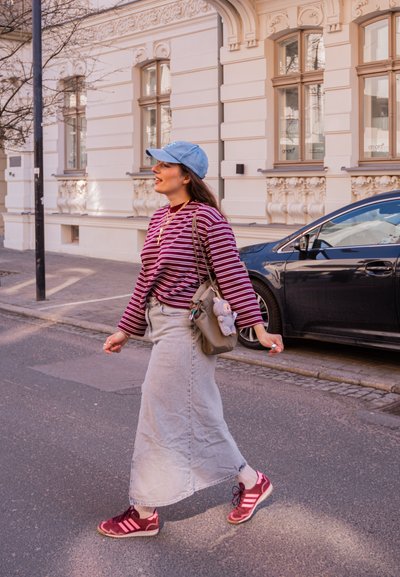 Mujer con camiseta de rayas, falda de mezclilla, gorra azul y zapatillas caminando por una calle de la ciudad junto a un coche negro estacionado y un edificio ornamentado.