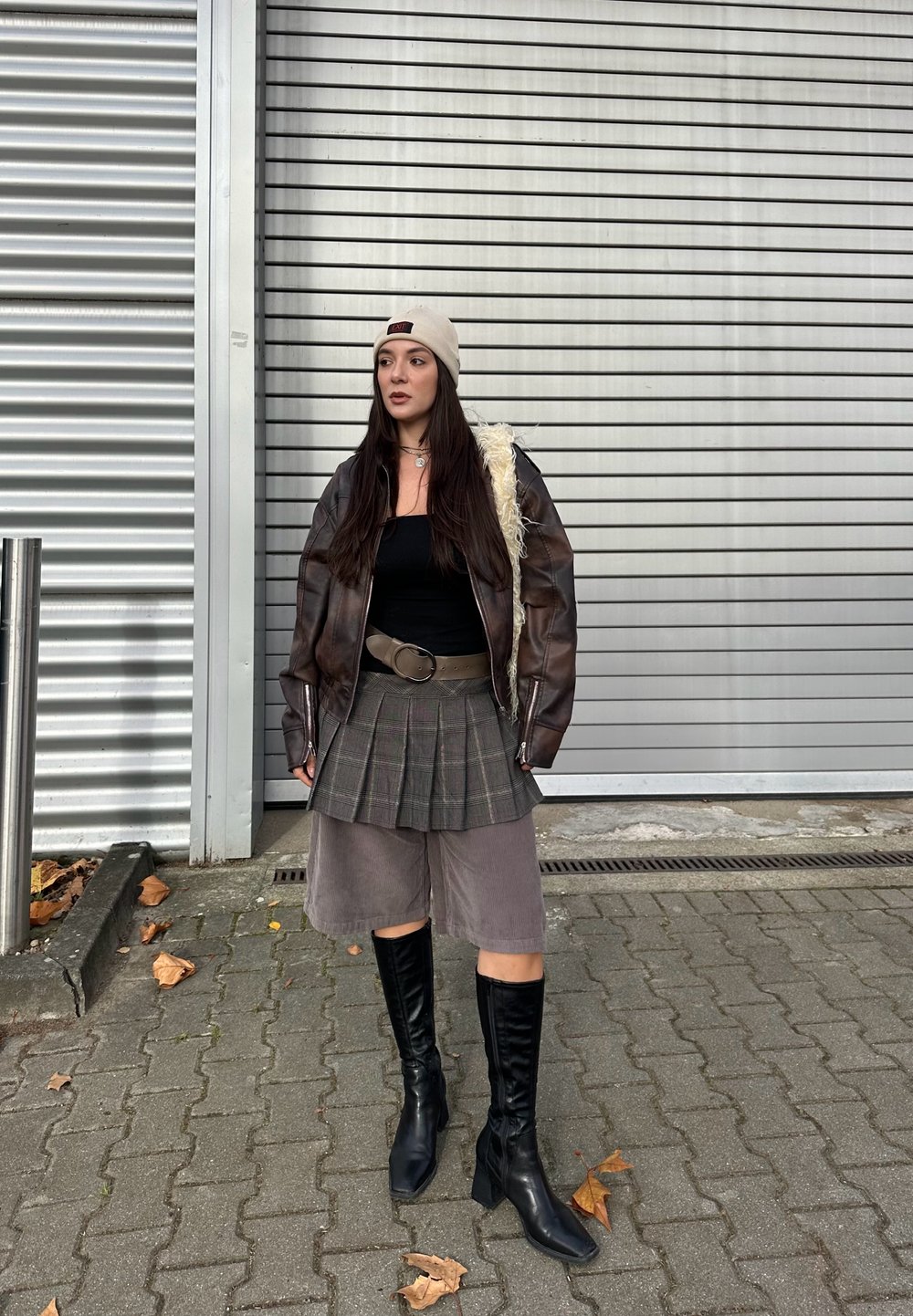 Rjava usnjena pilotska jakna z ovčjim krznom, črna majica, karirasta krila, sive kratke hlače in črni škornji do kolen, ki stojijo na pločniku.