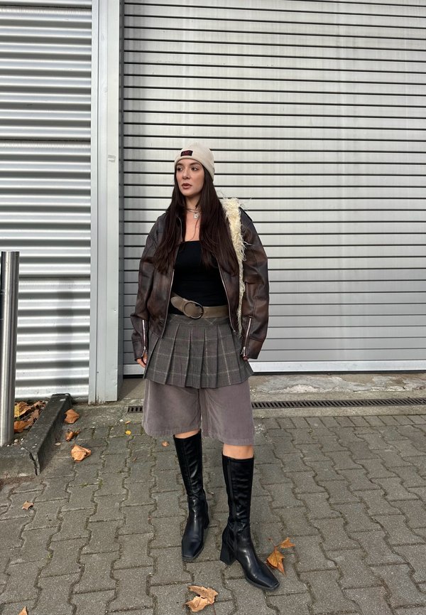 Brun läderbomberjacka med fårskinnsfoder, svart topp, rutig kjol, grå shorts och knähöga svarta stövlar som står på en trottoar.