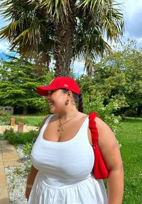 Κόκκινο καπέλο βάσης, άσπρο μπλουζάκι, κόκκινη τσάντα με ρυσετό λουρί, επιχρυσωμένα κολιέ σε στρώσεις. Το φόντο περιλαμβάνει παλμό και πράσινους θάμνους.