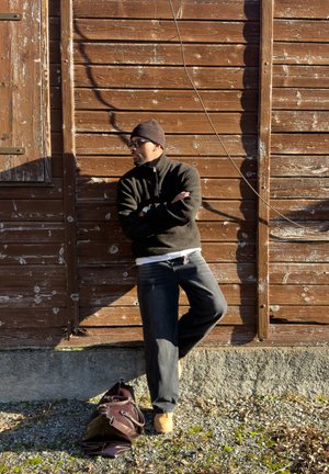 Hombre con gafas de sol, gorro y suéter negro apoyado contra una pared de madera desgastada con los brazos cruzados, bolsa en el suelo a su lado.