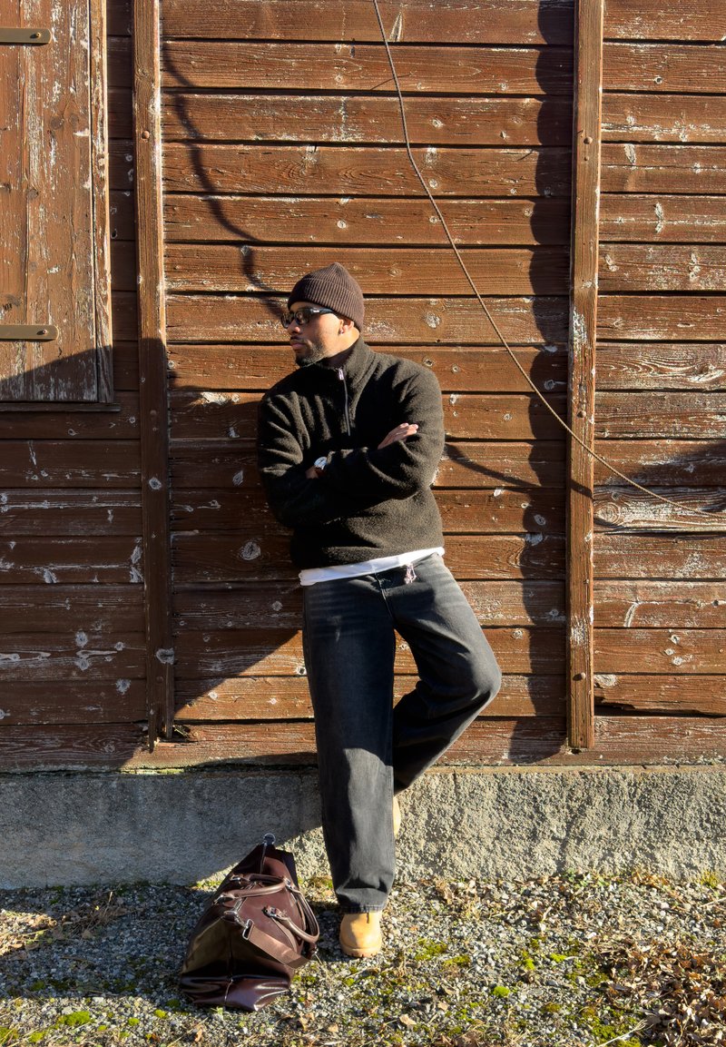 Homme portant des lunettes de soleil, un bonnet et un pull noir, appuyé contre un mur en bois vieilli, les bras croisés, avec un sac posé à côté de lui sur le sol.
