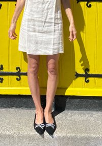 Witte linnen jurk, zwarte puntige schoenen met sprankelende lintaccenten, geposeerd voor een felgele deur. Soepele textuur, minimalistisch ontwerp.