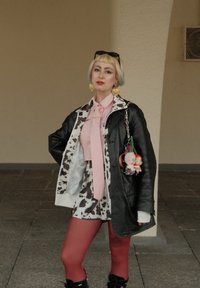 Persoon met kort blond haar en blauwe tips draagt een koeienprintsjas, een roze stropdas, rode panty, zwarte laarzen, hartoorbellen en houdt een tas met knuffeldieren vast.