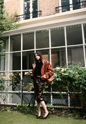 Jachetă din piele maro, top negru și fustă creion bordo. Tocuri stiletto. Foliaj verde și feronerie mari în fundal.