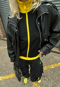Czarna skórzana kurtka motocyklowa na czarnej i żółtej bluzie na zamek, do tego czarne spodnie cargo i żółte sneakersy. Zawiera zamki i elementy metalowe.