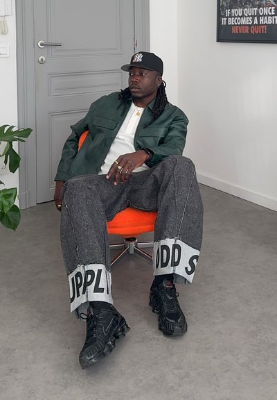 Hombre con gorra de béisbol, chaqueta verde y pantalones anchos con texto, sentado relajado en una silla giratoria naranja en una habitación gris con un cartel motivacional.