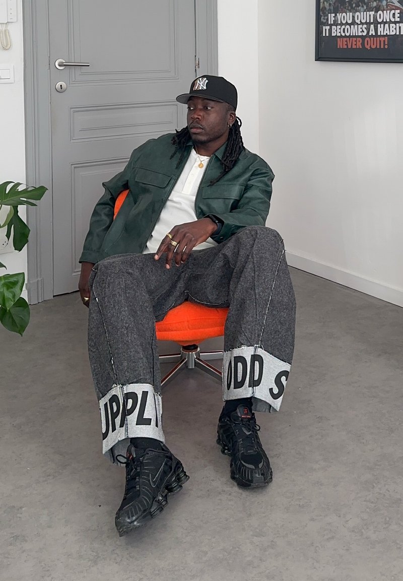 Mężczyzna w czapce z daszkiem, zielonej kurtce i szerokich spodniach z tekstem siedzi zrelaksowany na pomarańczowym obrotowym krześle w szarym pokoju z motywacyjnym plakatem.