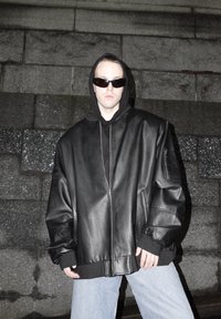 Personne portant une veste en cuir noire surdimensionnée, un sweat à capuche noir avec la capuche relevée, des lunettes de soleil noires et un jean bleu clair, debout contre un mur en pierre.