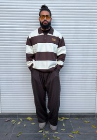 Partainen mies nutturalla, jolla on suuret keltaiset aurinkolasit, ruskeavalkoraidallinen neule ja väljät tummat housut, seisoo valkoisen rullakaihtimen edessä.