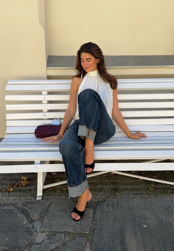 Mujer con camiseta blanca sin mangas y vaqueros remangados sentada con las piernas cruzadas en un banco blanco al aire libre con un bolso pequeño morado a su lado.