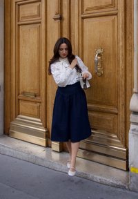 Vrouw in een witte blouse en marineblauwe rok die voor grote, sierlijke houten deuren staat en een zonnebril en een witte handtas vasthoudt.