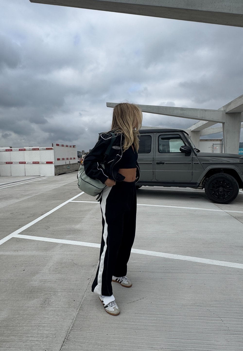 Sort-hvide træningsbukser, sort cropped top og en let sort jakke. Bærer en lysegrøn taske. Grå SUV parkeret i nærheden.