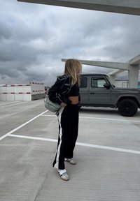 Sort-hvide træningsbukser, sort cropped top og en let sort jakke. Bærer en lysegrøn taske. Grå SUV parkeret i nærheden.