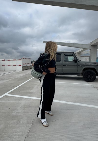 Pantalones deportivos en blanco y negro, top negro recortado y chaqueta negra ligera. Lleva una bolsa verde claro. SUV gris estacionado cerca.