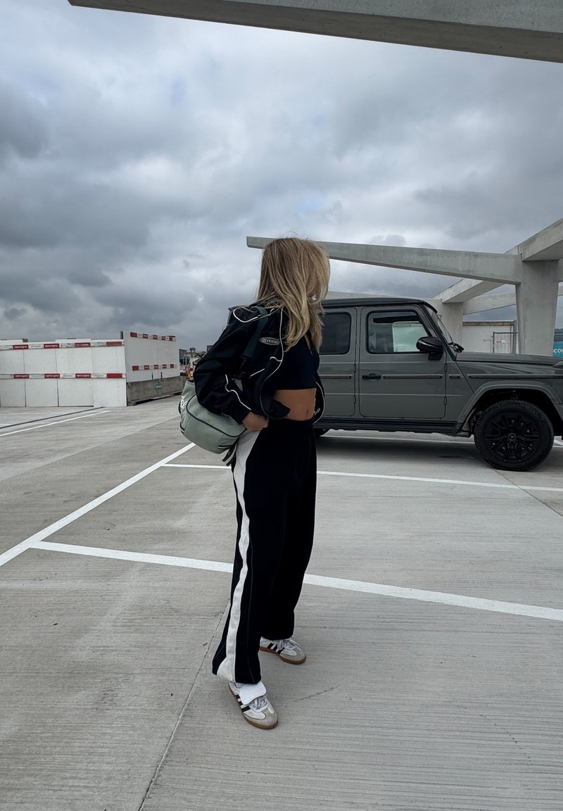 Pantalon de survêtement noir et blanc, haut noir court et une veste noire légère. Porte un sac vert clair. Un SUV gris est garé à proximité.