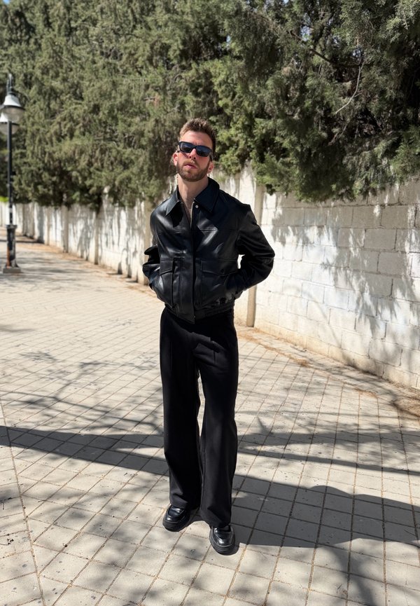 Mężczyzna w czarnych okularach przeciwsłonecznych, czarnej skórzanej kurtce i czarnych spodniach stoi na oświetlonym słońcem chodniku, za nim drzewa i biała ceglasta ściana.
